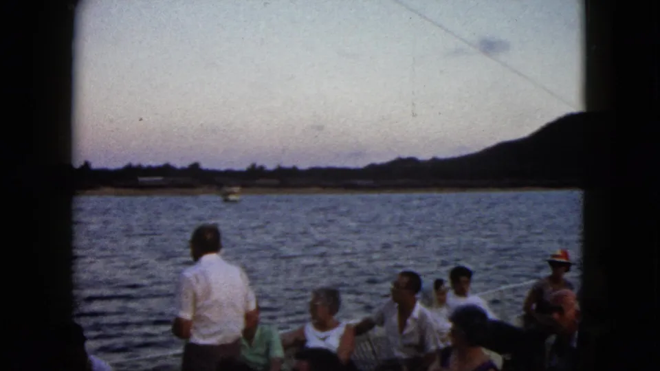 1962 Acapulco home-movie frame showing passengers on a tour boat looking toward water, shoreline, and hills in fading light.