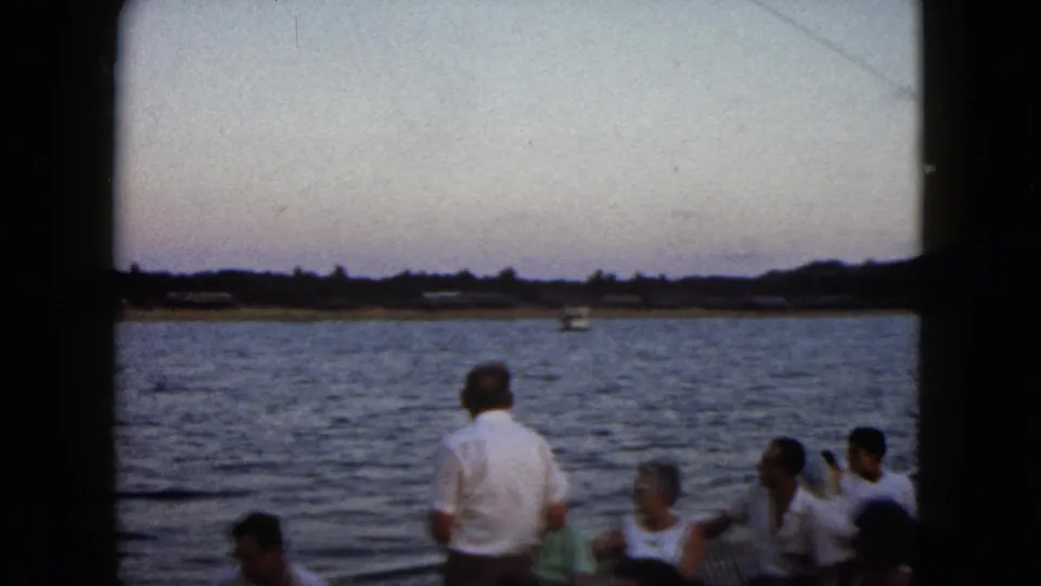 1962 Acapulco frame with tourists seated and standing on a boat, seen against calm water, a distant shore, and dusky sky.
