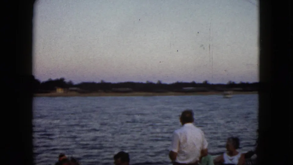 Home-movie view from a 1962 Acapulco boat, with tourists partly framed by dark film edges and shoreline beyond the water.