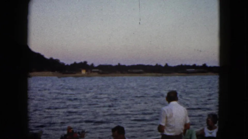 1962 Acapulco home-movie frame showing a cropped group of boat passengers, rippled water, low shoreline, and dark hillside at dusk.