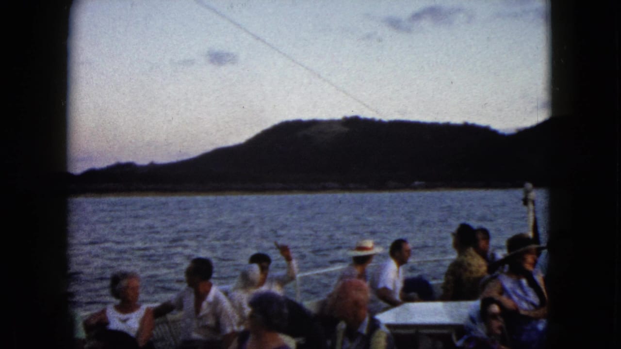 Seated adults on a tourist boat in Acapulco, Mexico, seen against calm water, a dark hillside, and a pale evening sky.