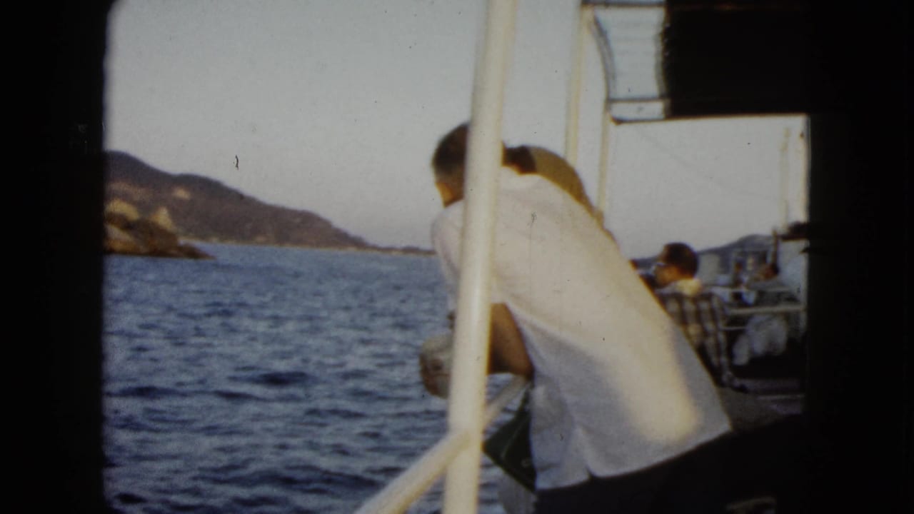 Man in a white shirt leans on a boat railing beside the sea and distant coastline.