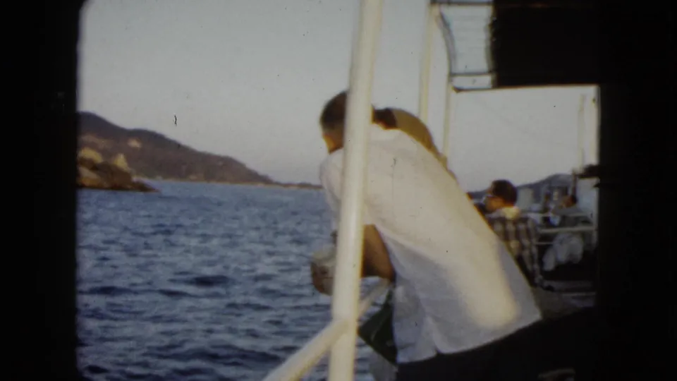 1962 Acapulco home-movie frame showing a man in a white shirt leaning on a boat railing with sea and mountains beyond.