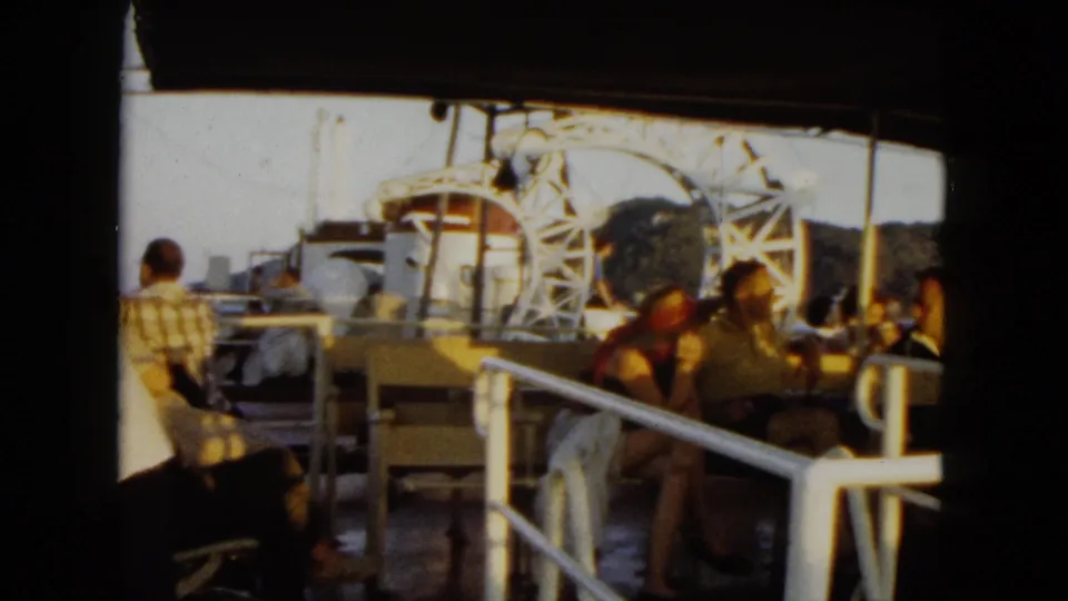 1962 Acapulco home-movie frame showing tourists seated on a boat deck beneath a canopy with mountains behind.