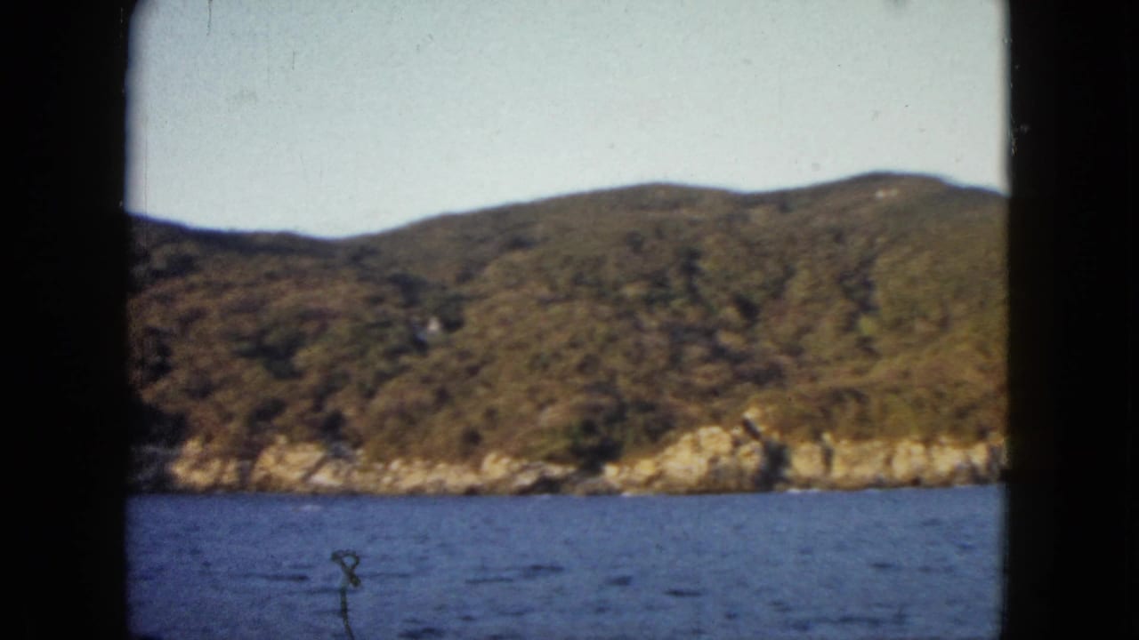 1962 Acapulco home-movie frame showing blue water and a rugged green coastal hillside from a boat viewpoint.