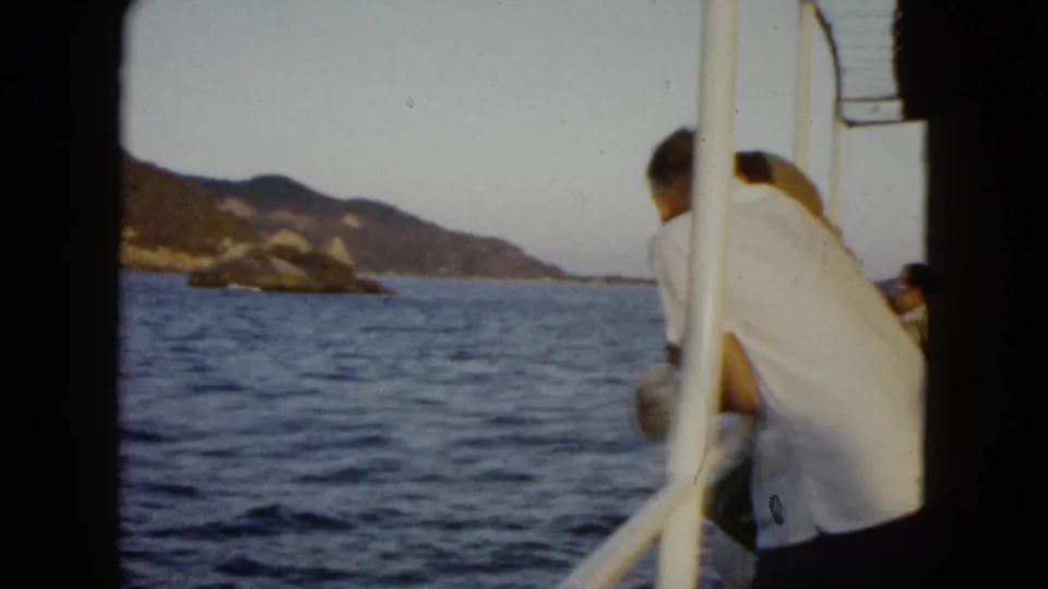A passenger on a boat in Acapulco, 1962, leans toward the rail as blue water and rocky green coastline fill the background.