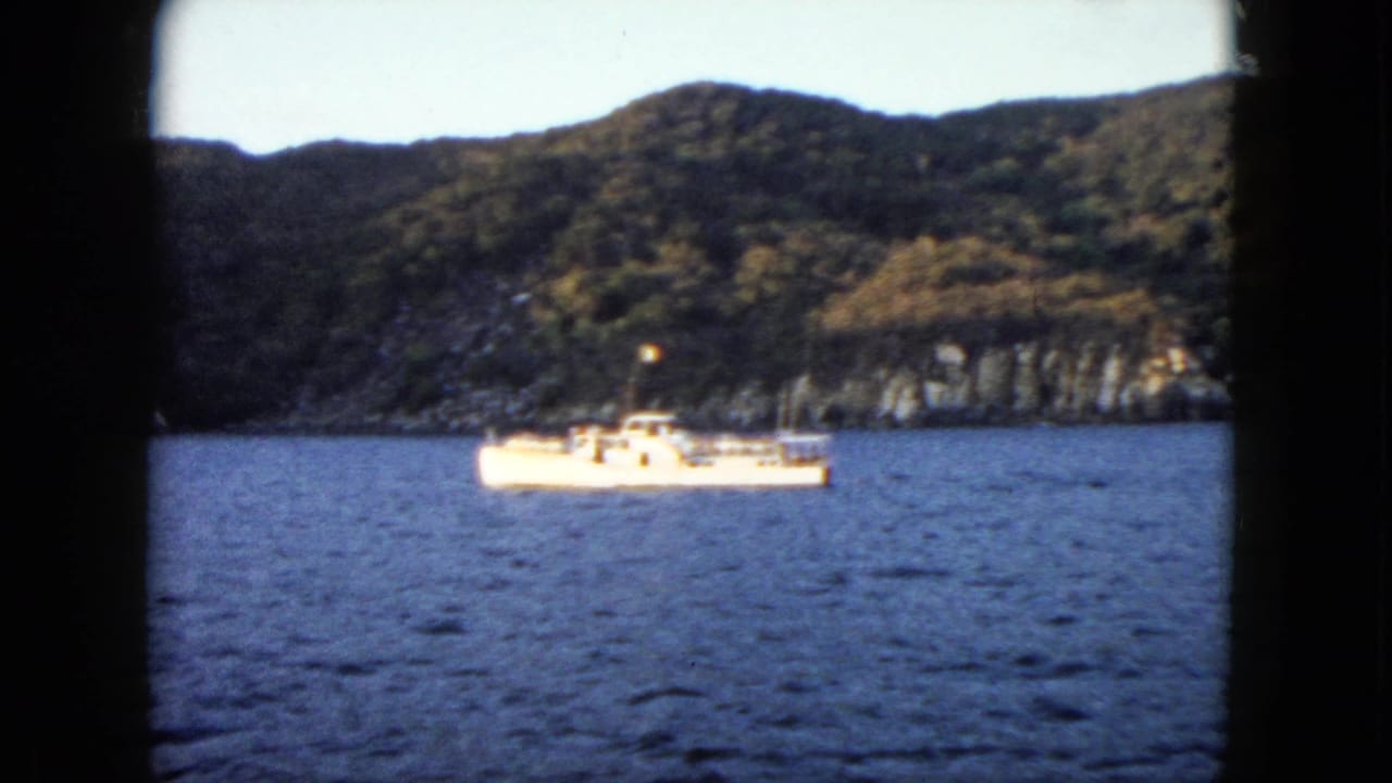 1962 Acapulco home-movie frame showing a small white boat crossing blue bay water in front of wooded hills and a rocky coast.