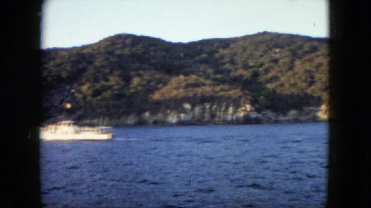 White boat near the left edge with blue water, wooded hills, and rocky coast.