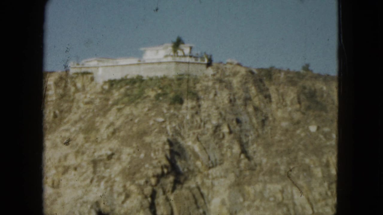 1962 Acapulco home-movie frame showing a white cliff-top building above rugged rock and sparse greenery.