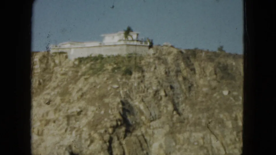 1962 Acapulco home-movie frame showing a white cliff-top building above rugged rock and sparse greenery.