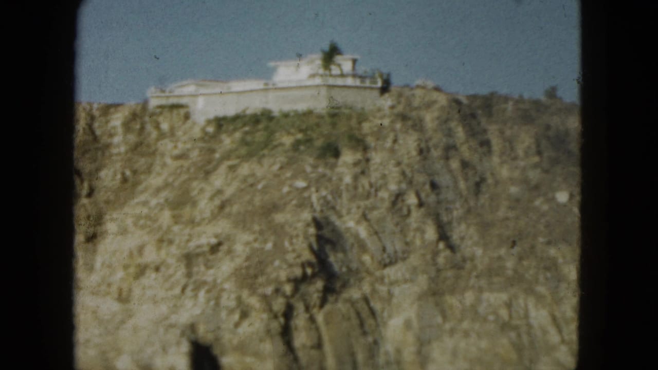 1962 Acapulco view of a white structure perched on a rocky cliff, framed by 8mm gate edges and open sky.