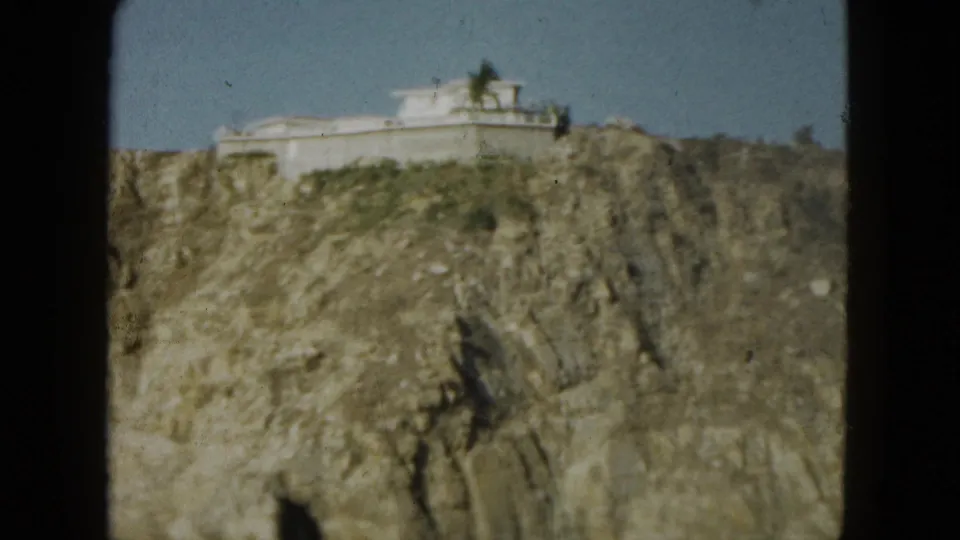 1962 Acapulco view of a white structure perched on a rocky cliff, framed by 8mm gate edges and open sky.