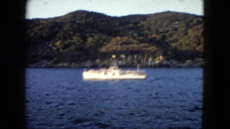 1962 Acapulco frame showing a small boat crossing blue water below green hills and rocky shoreline.