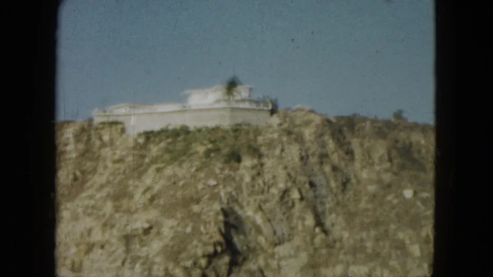 1962 Acapulco coastal frame showing a pale clifftop building above a rocky hillside under clear sky.