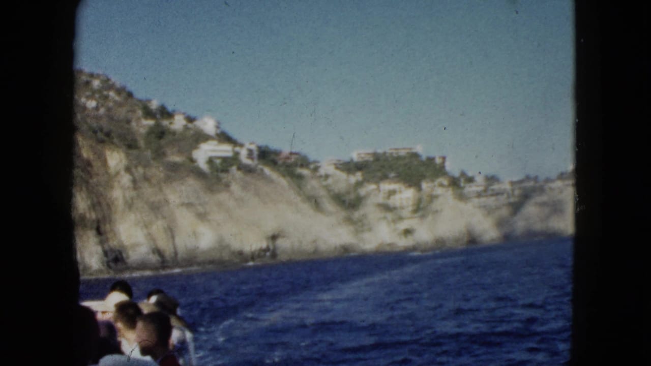 Boat passengers look toward rocky coastal cliffs and blue water under a clear sky.