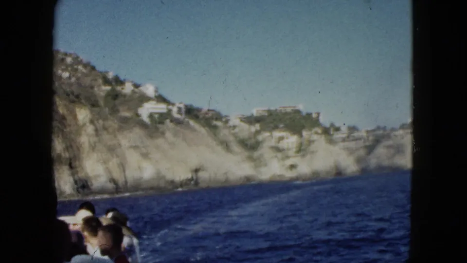 1962 Acapulco home-movie view from a boat, with passengers in the foreground and rocky cliffs with white buildings beyond the water.