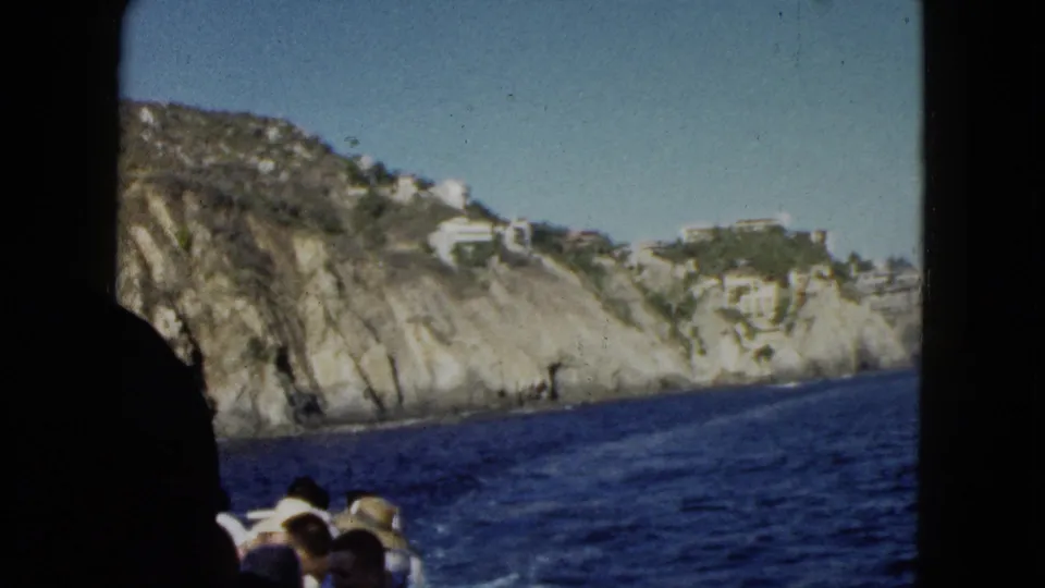 Acapulco coastline filmed from inside a moving boat, with a dark vertical frame edge, seated passengers, cliffs, and hillside buildings visible.