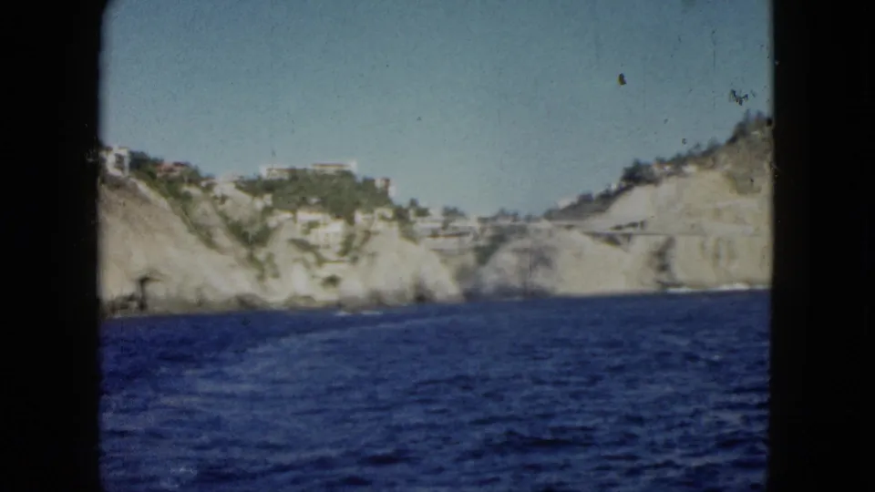 1962 home-movie view of Acapulco's cliffside coast, with blue water, pale rock, and hillside buildings.