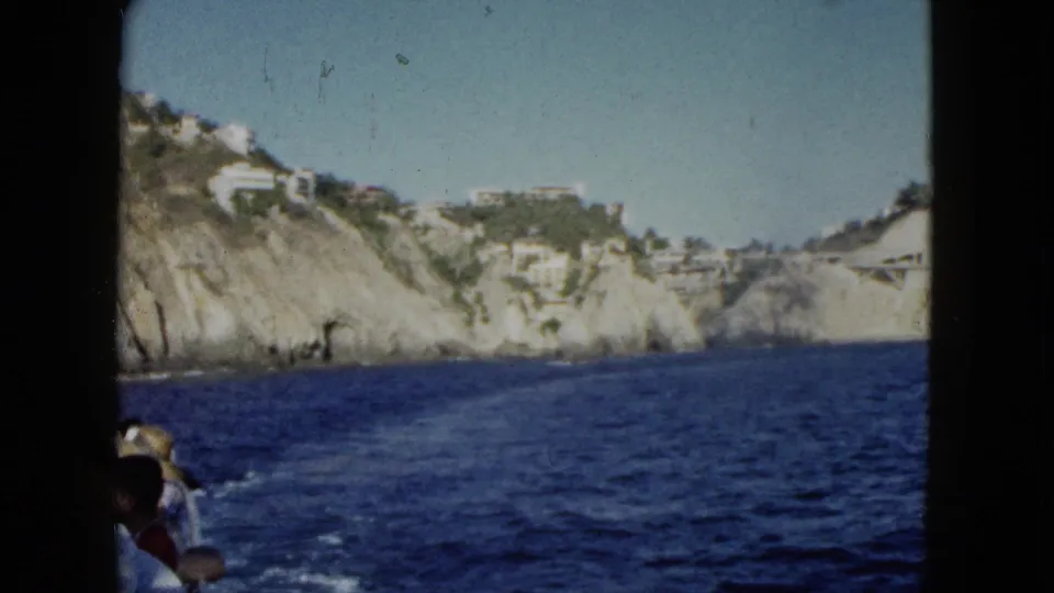 Final 1962 Acapulco boat view with passengers at the lower edge, ocean wake, cliffs, and hillside buildings.