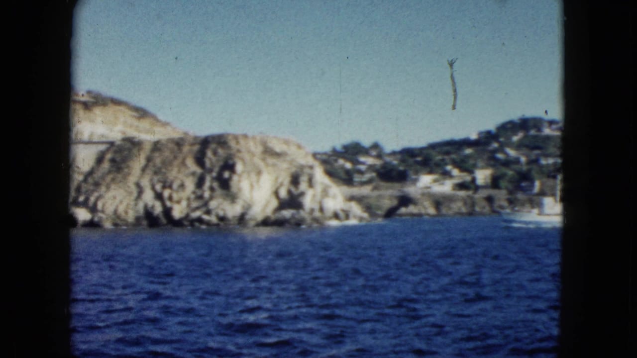 1962 8mm home-movie frame from Acapulco showing pale rocky cliffs, deep blue water, scattered hillside buildings, and visible film-gate edges.