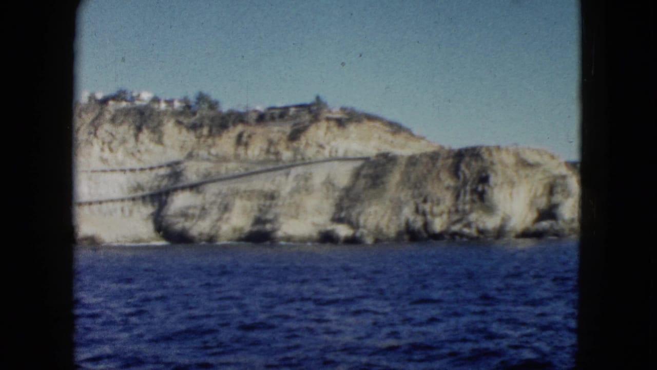 1962 Acapulco coastal view with rugged cliffs, a long road or bridge crossing the rock face, hillside structures, and blue water in the foreground.