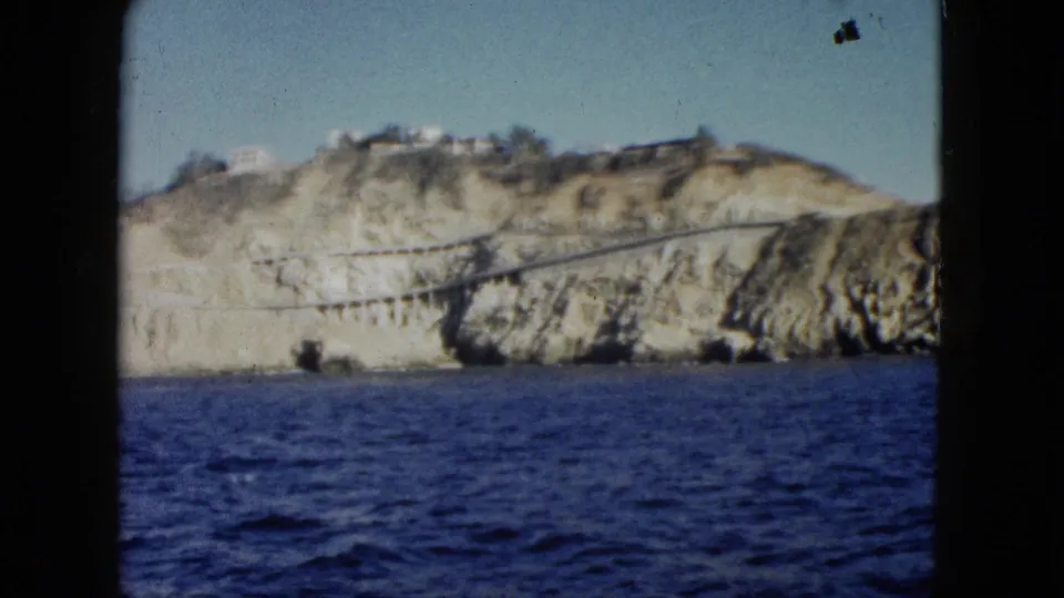 Soft 1962 8mm frame showing Acapulco cliffs above blue water, with hillside buildings and a road or bridge visible across the rocky coastline.