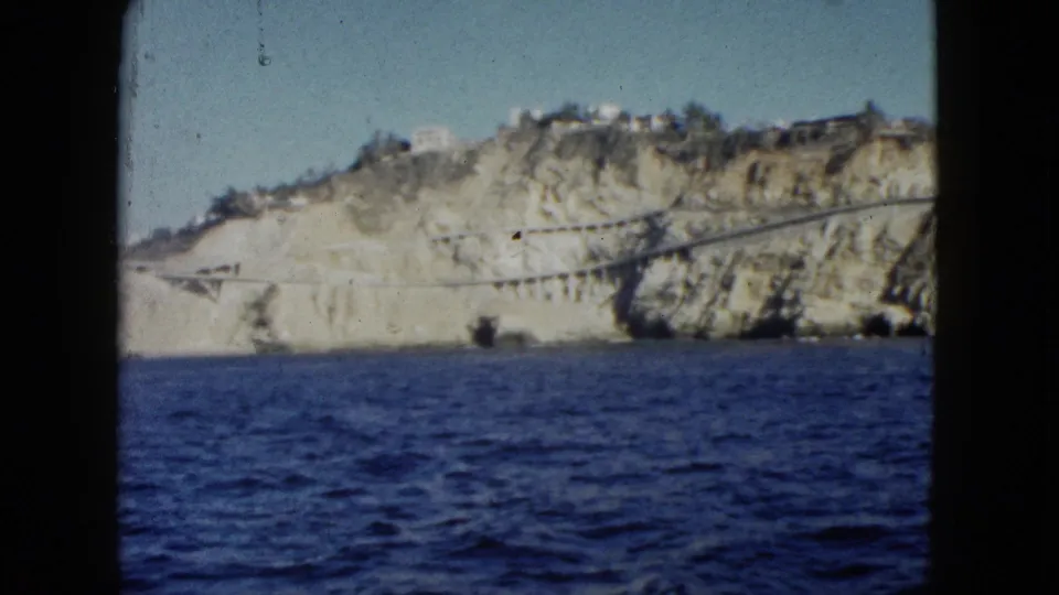 1962 Acapulco home-movie frame with a wider look at blue water, pale rocky cliffs, hillside buildings, and the road or bridge line along the coast.