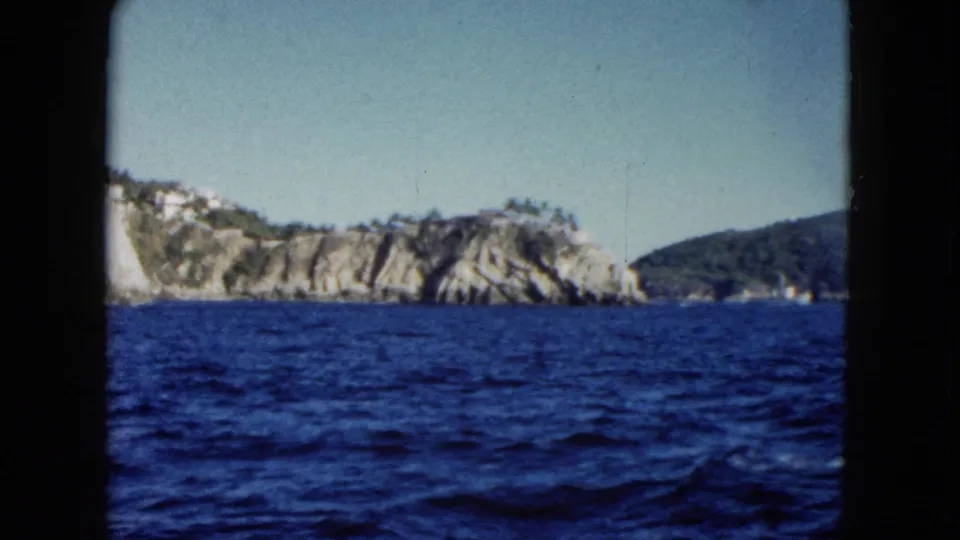 1962 8mm view from the water toward rocky Acapulco cliffs, distant white hillside buildings, and a clear sky framed by dark film borders.