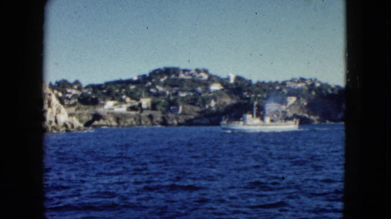 Small white boat near rocky shoreline and hillside houses across dark blue water.