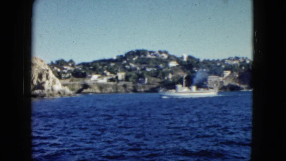 Visitor-shot 8mm frame of a white boat crossing blue water near Acapulco's rocky coast and hillside buildings in 1962.