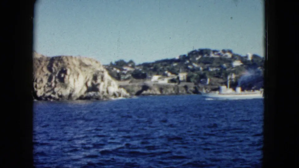 1962 Acapulco home-movie frame showing choppy blue water, rocky cliffs, distant hillside buildings, and a small white boat near shore.