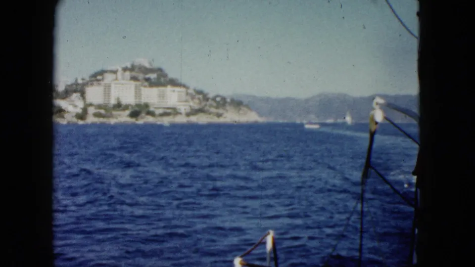 1962 Acapulco 8mm frame showing a boat-view approach to blue water and distant hillside resort buildings.