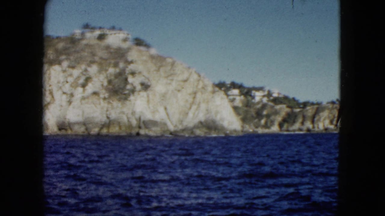 Rocky coastal headland above blue water, framed by dark vertical film edges.