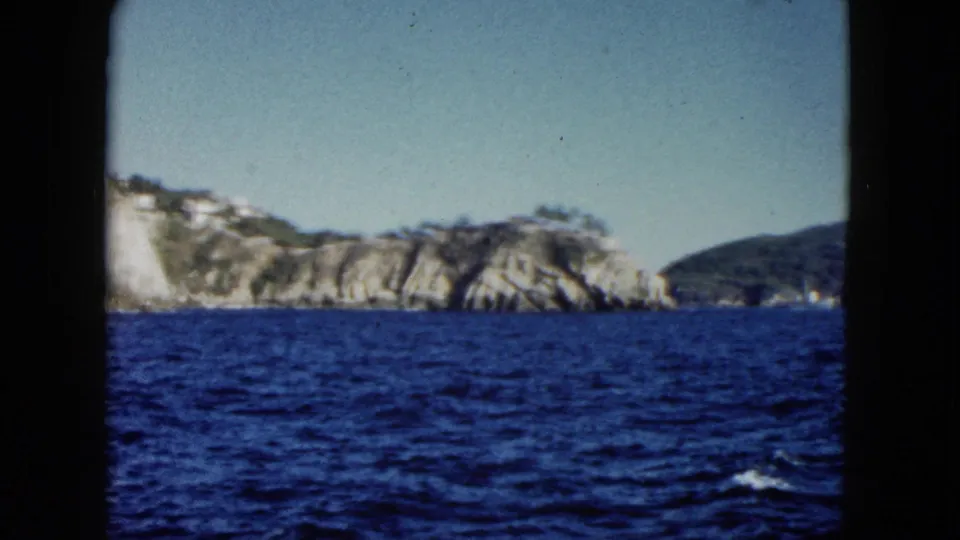 1962 Acapulco 8mm frame showing rocky coastal cliffs, distant hills, and open water from a boat.