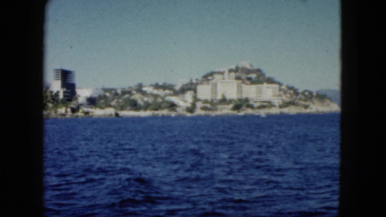 1962 Acapulco home-movie frame showing deep blue water and a distant hillside with white resort buildings under a pale sky.