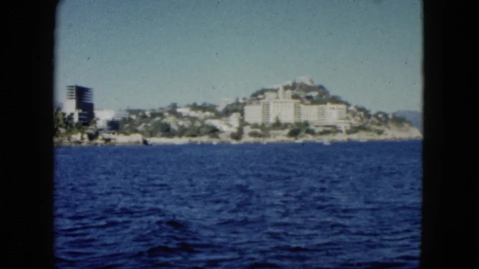 1962 Acapulco home-movie frame showing deep blue water and a distant hillside with white resort buildings under a pale sky.
