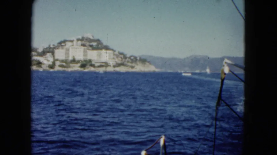 1962 Acapulco boat-view frame with ocean, hillside buildings, distant mountains, and a small rail at the edge of the shot.