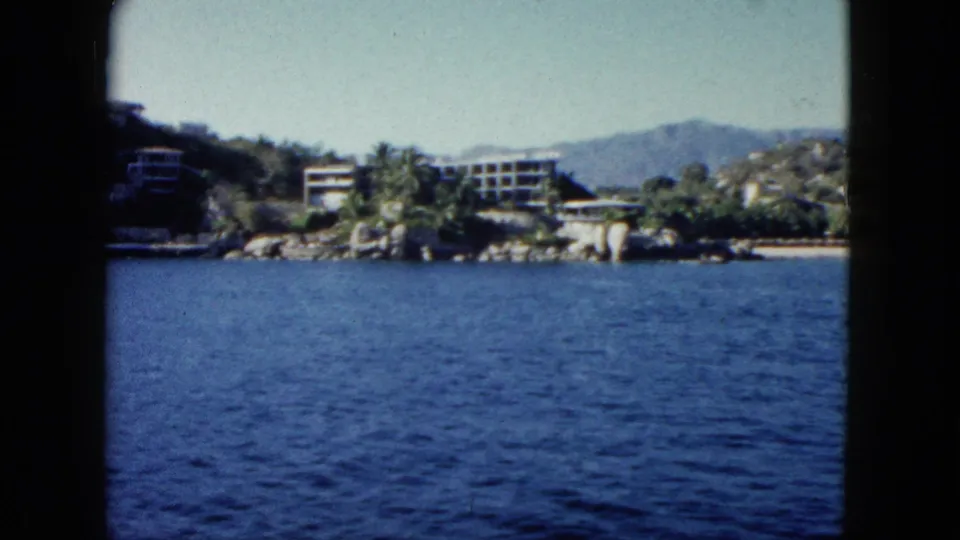 1962 Acapulco home-movie frame looking from a boat across rippling water toward a tropical shoreline with buildings and hills.
