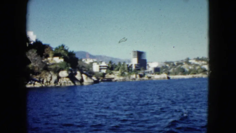 1962 Acapulco shoreline seen from a moving boat, with a rocky point, palms, hillside buildings, and a tall coastal tower.