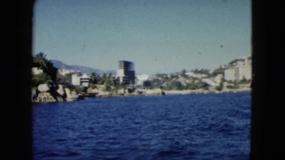Acapulco coastal scene from 1962 showing bay water, a rocky shore, palms, a tall building, and hillside resort structures.