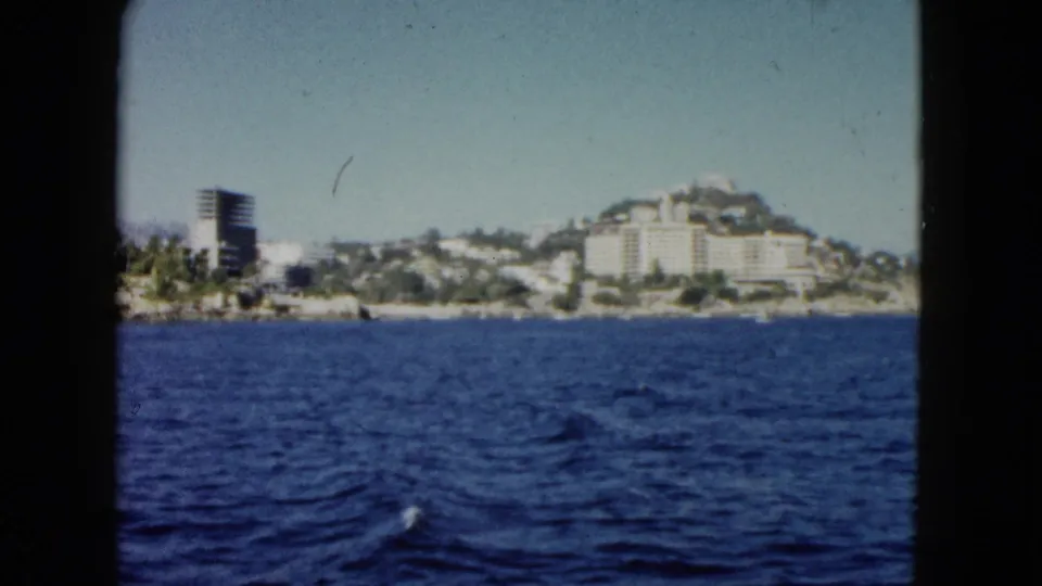 Wide 1962 Acapulco boat view across deep blue water toward hillside hotels or apartments and a dark tower on the coast.