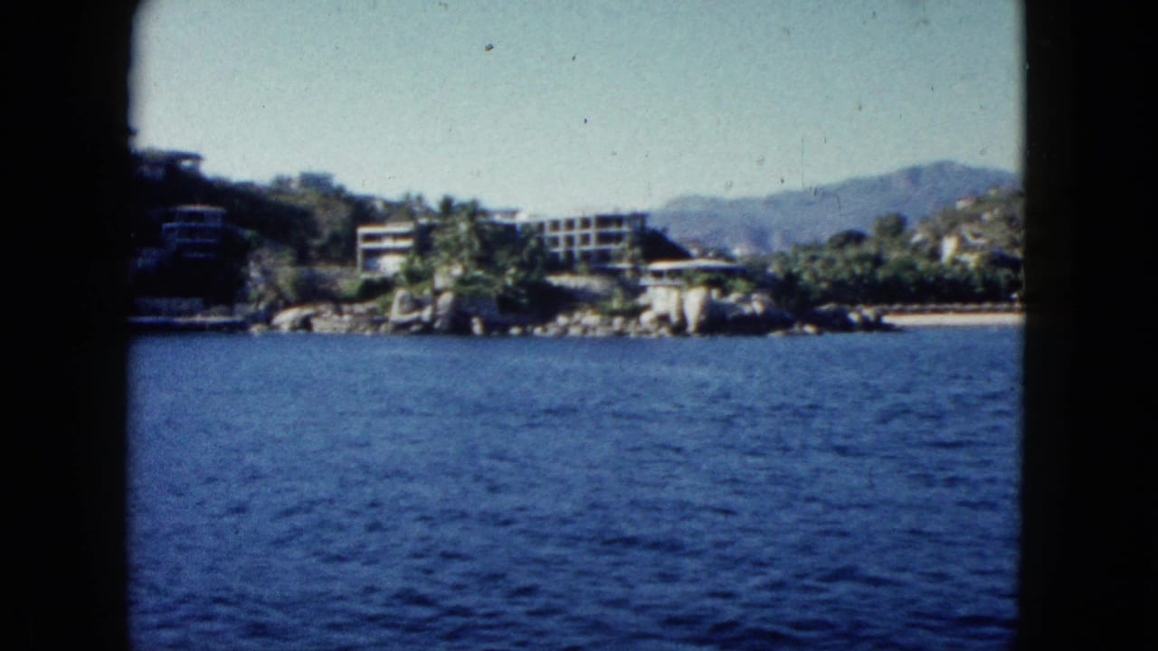 1962 Acapulco boat-view frame showing bay water, rocky shore, palm trees, low modern buildings, distant hills, and dark edge framing.
