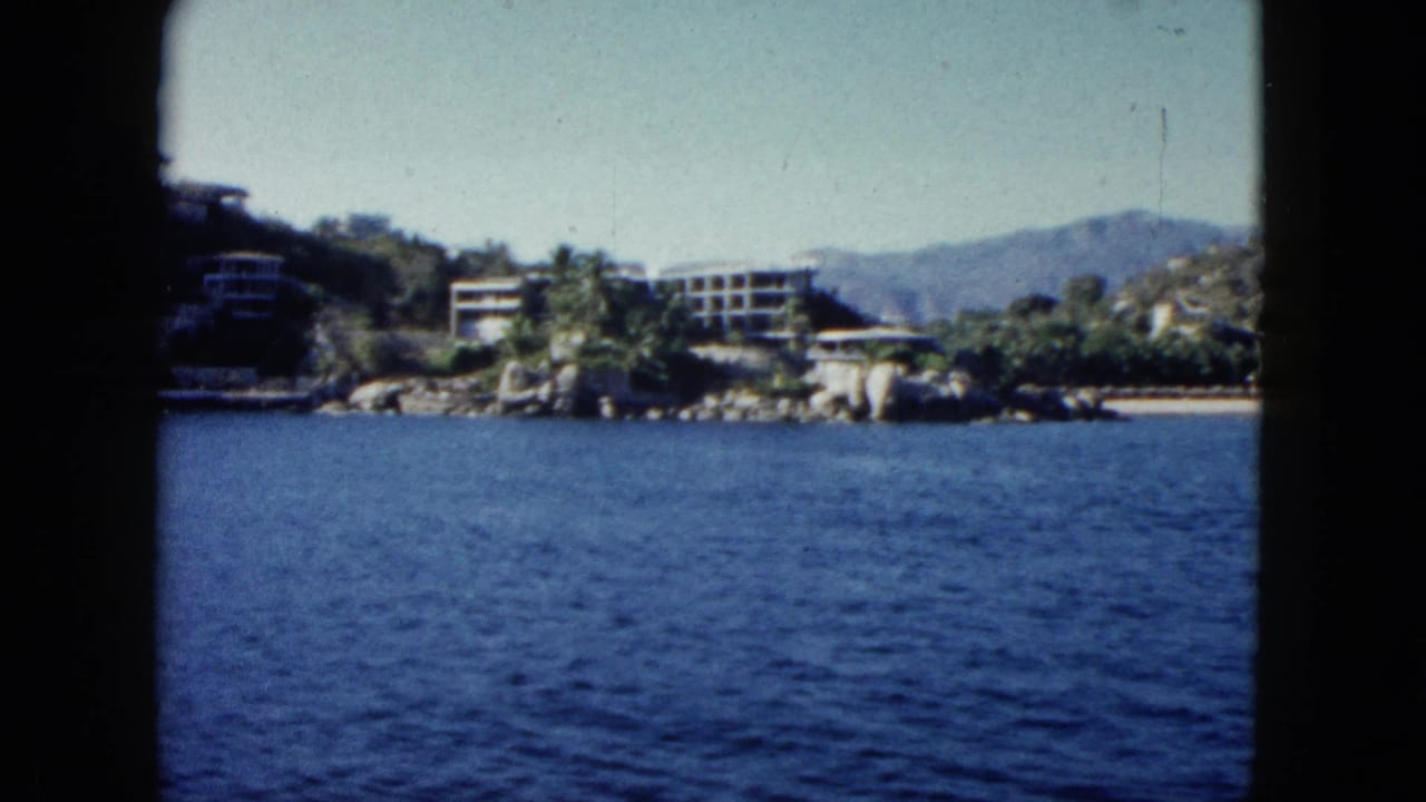 Waterfront buildings and palms sit along a rocky shore beyond rippling blue water and dark side framing.