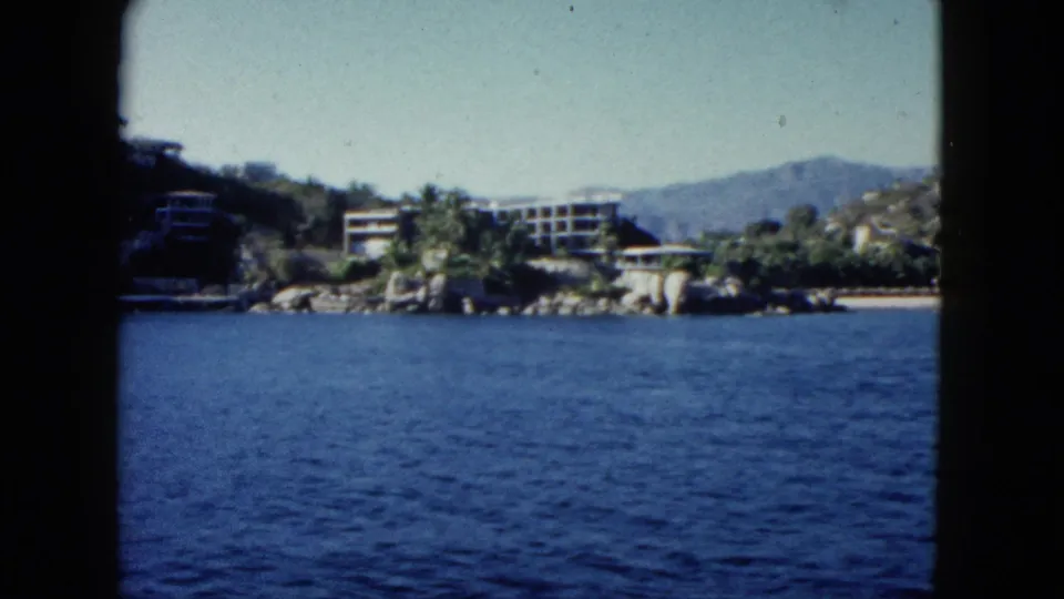1962 Acapulco shoreline view from water, showing blue bay, rocks, palms, low modern structures, distant mountains, and soft 8mm color.