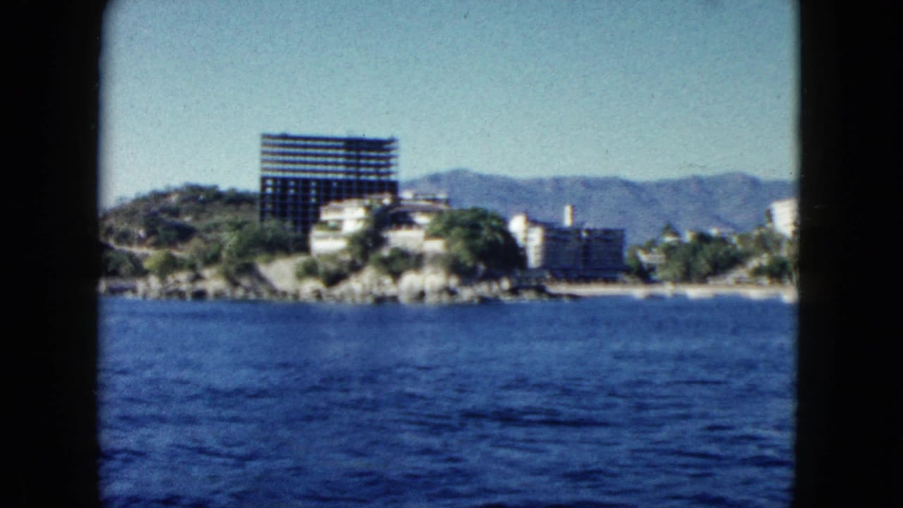 1962 Acapulco home-movie frame from the water, showing blue ocean, rocky shore, hotels, a dark high-rise, green hills, and mountains.