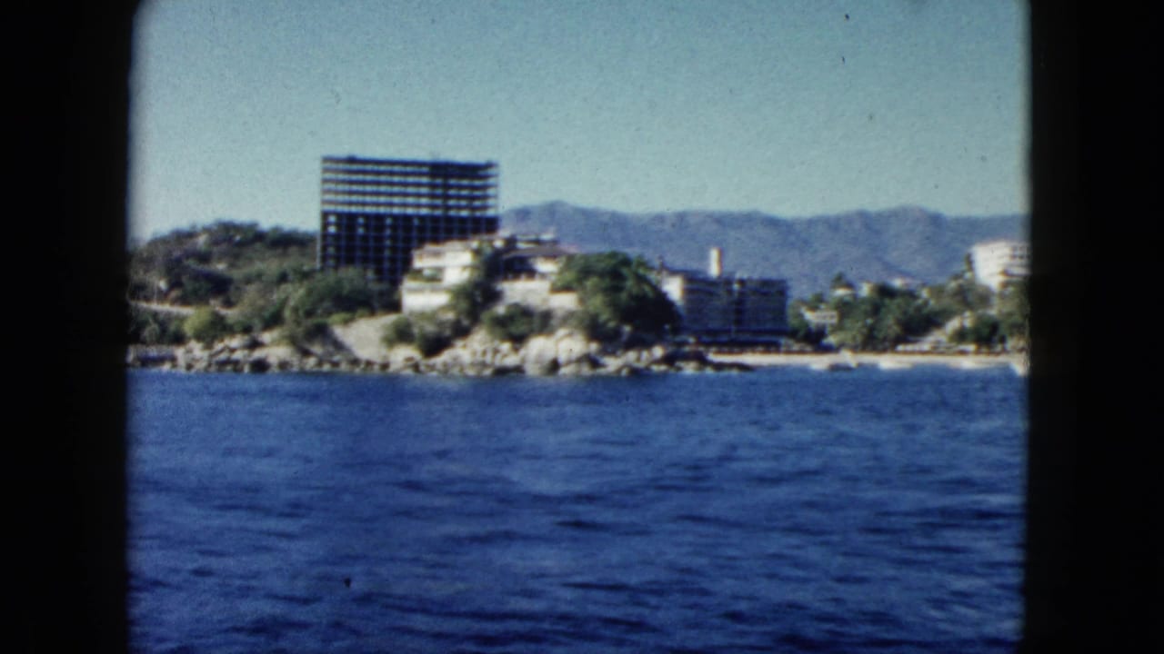 Ocean view toward Acapulco hotels, a dark high-rise structure, rocky shore, and mountains.