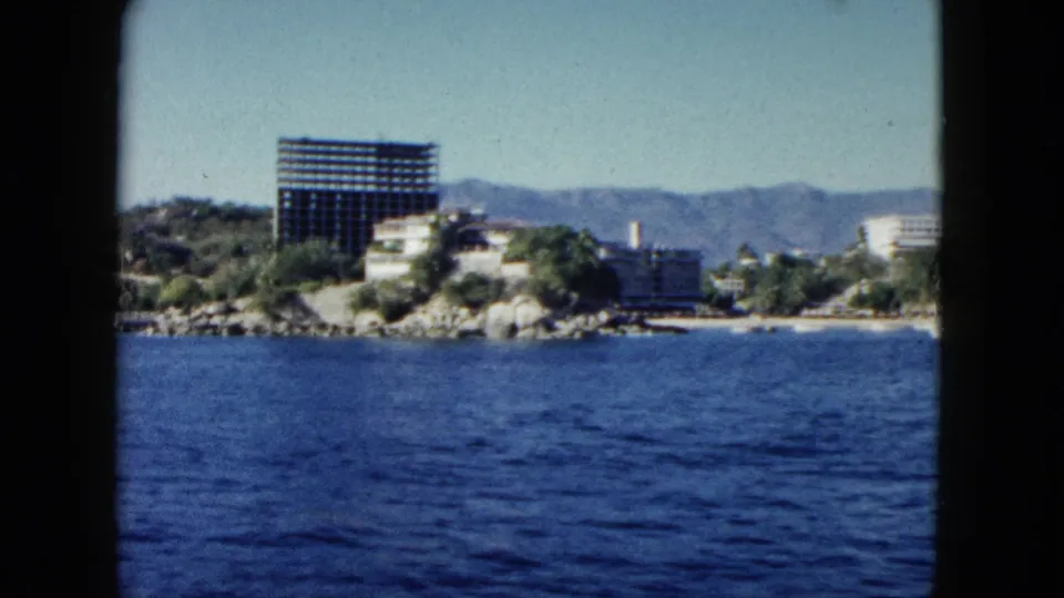1962 home-movie view of Acapulco from the ocean, showing shoreline buildings, rocks, vegetation, a prominent high-rise, and distant mountains.