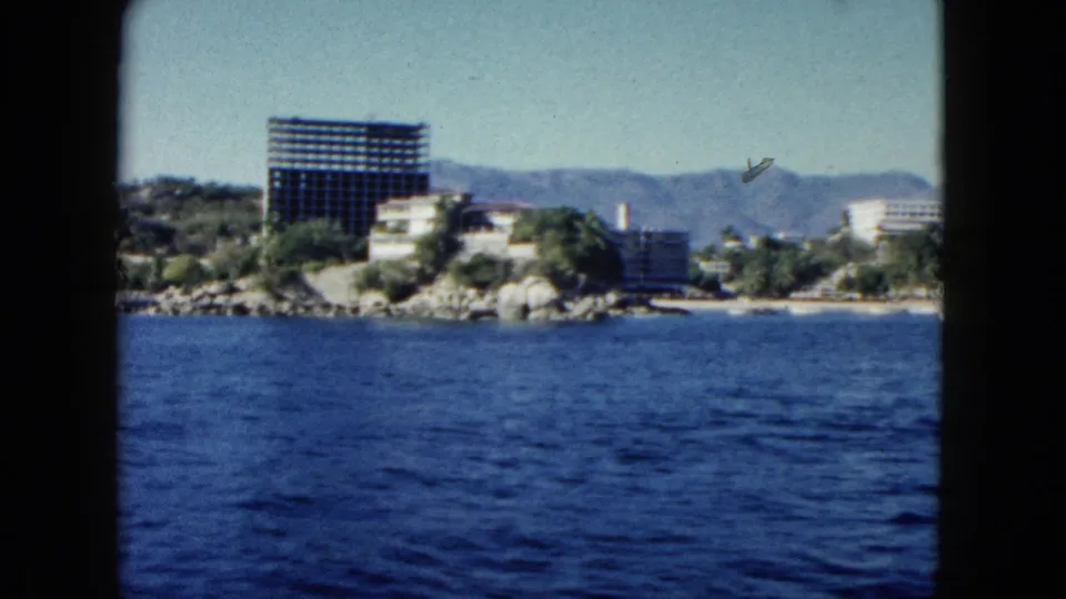 1962 Acapulco boat-view frame with ocean foreground, hotel-lined shore, high-rise construction, hillside greenery, mountains, and visible 8mm film wear.