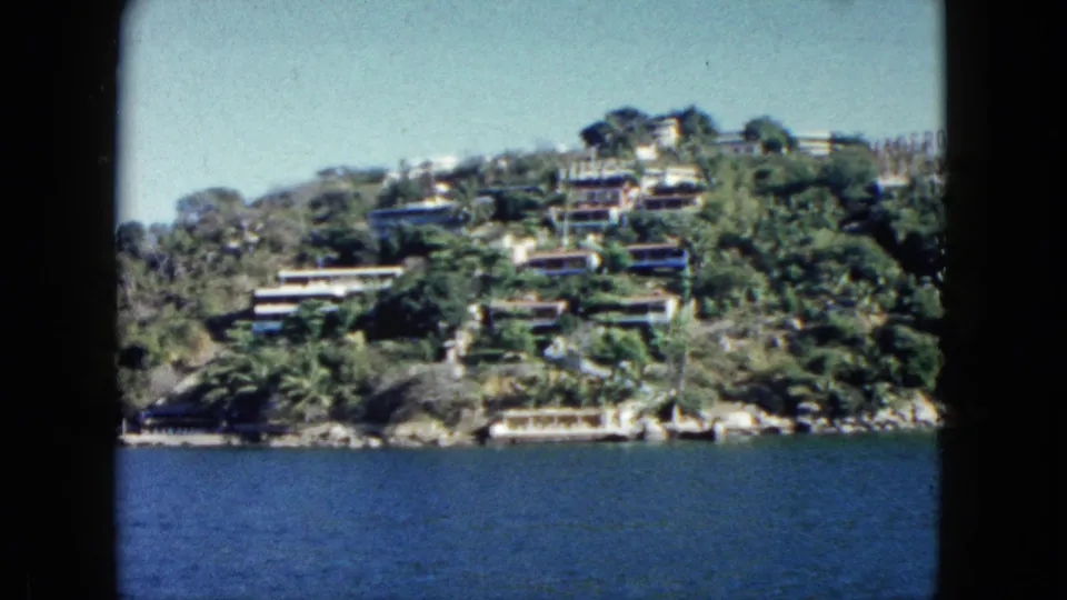 Boat-level 8mm view of Acapulco hillside housing and shoreline vegetation, with blue water across the lower frame.