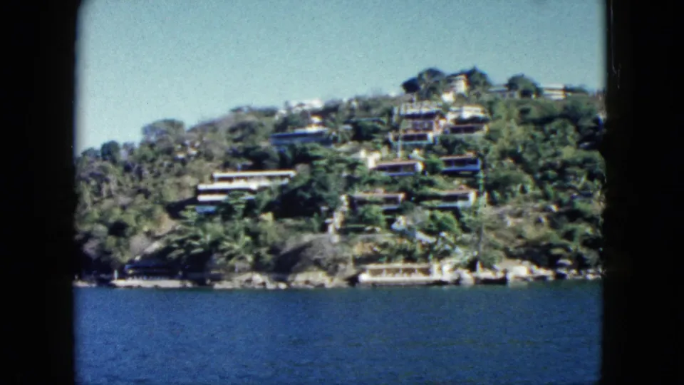 1962 Acapulco shoreline view with hillside homes, dense vegetation, and a tall structure visible above the water.
